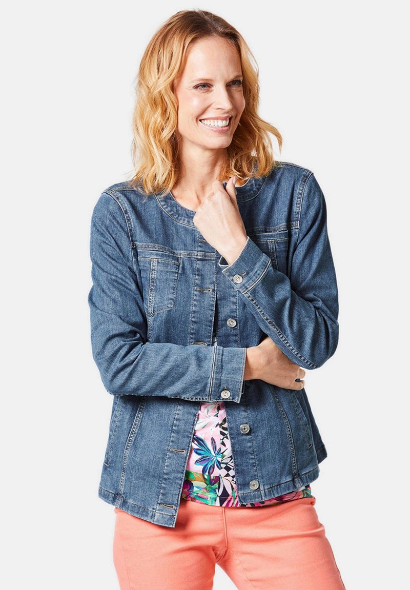 Blaue Jeansjacke mit rundem Halsausschnitt, Knopfverschluss und zwei Brusttaschen, getragen über einem mit Blumenmuster bedruckten Oberteil und rosa Hosen.