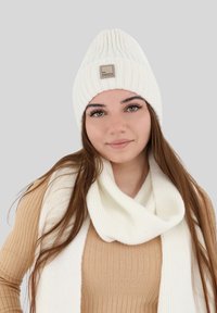 Bonnet en tricot côtelé blanc avec une patch logo, associé à une écharpe en tricot côtelé blanc assortie. Les deux articles présentent une texture douce et un design cintré.