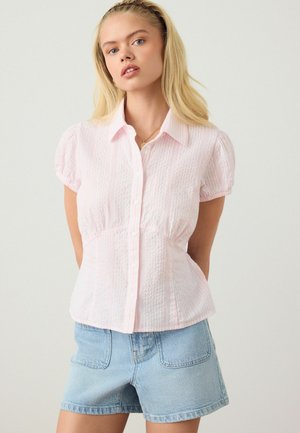 Young woman with long blonde hair wearing a light pink textured short-sleeve button-up blouse and light blue denim shorts.