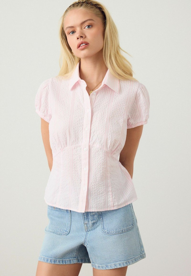 Jeune femme aux longs cheveux blonds portant un chemisier texturé rose clair à manches courtes boutonné et un short en jean bleu clair.