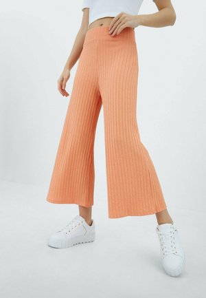 Vrouw draagt een hooggetailleerde, wijde, geribbelde perzikkleurige broek en witte plateau sneakers, staand tegen een effen witte achtergrond.