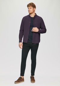Dunkel-lila Hemd mit Knöpfen über einem schwarzen Rundhalsausschnittshirt, kombiniert mit schwarzen, schmalen Hosen und braunen Sneakern mit strukturierter Oberfläche.