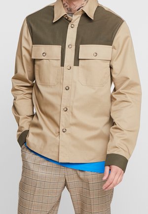 Mann trägt beige-olivgrünes Hemd mit Brusttaschen über blauem Hemd und beigen karierten Hosen vor einfarbigem Hintergrund.