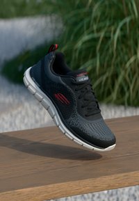 Black athletic shoe with a lightweight mesh upper, textured design, and white sole. Features red accents and a logo on the side.