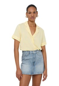 Camicia gialla a maniche corte, senza colletto e dalla vestibilità rilassata, abbinata a una minigonna in denim azzurro chiaro. Tessuto dalla texture liscia.