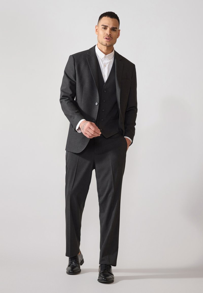 Dunkelgrauer Dreiteiler mit Schlangenlapel, passender Weste und geraden Hosen. Darunter ein weißes Hemd, ergänzt durch schwarze Schuhe.