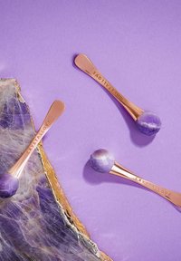 Tre rulli per il viso in giada viola con manici in oro rosa, che presentano una superficie liscia e lucida, su uno sfondo lavanda con una fetta di cristallo.
