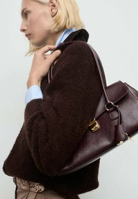 Bordeaux leren handtas met gouden hardware, strak ontwerp en een geprofileerde afwerking, vastgehouden tegen een donkerbruine fuzzy trui.