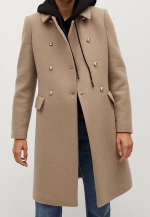 Manteau en laine beige à double boutonnage sur un sweat à capuche noir avec cordons, porté avec un jean denim bleu, ne montrant que les mains et le torse.