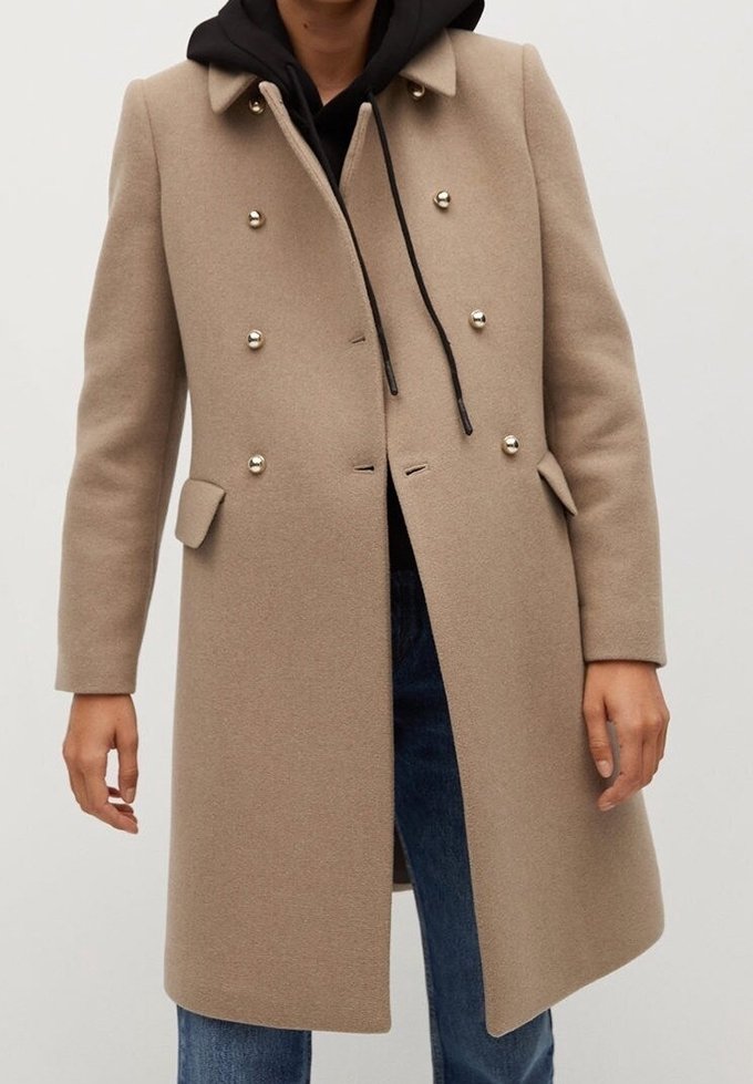Manteau en laine beige à double boutonnage sur un sweat à capuche noir avec cordons, porté avec un jean denim bleu, ne montrant que les mains et le torse.