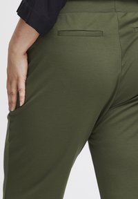 Olivgröna byxor gjorda av stretchmaterial, med en smooth yta och två bakfickor, där en hand vilar på sömmen.