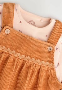 Salopette en velours côtelé orange avec des bords en scallop, accents de boutons en bois, superposée sur un haut rose clair avec un motif floral.