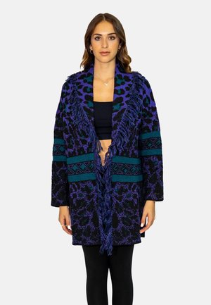 Cardigan lavorato a maglia in viola e nero con stampa leopardata, righe turchesi, colletto frangiato e design aperto, indossato sopra un top nero.