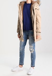 Giacca puffer beige con cappuccio foderato in pelliccia, maglione blu navy, jeans blu strappati con decorazioni e sneakers bianche.