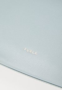 Világoskék bőr táska texturált felülettel, hangsúlyos arany "FURLA" logóval. Egyszerű, téglalap alakú, sima élekkel.