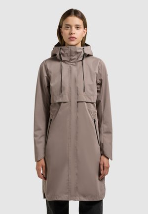 Nő hosszú taupe színű kapucnis esőkabátban, fekete cipzáras zsebekkel és zsinórokkal, egyszínű világosszürke háttér előtt áll.