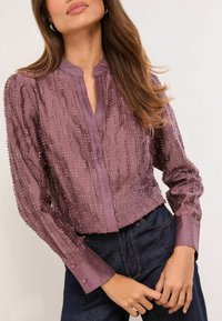 Lila hosszú ujjú blúz texturált anyagból, finom bordázattal és gallér nélküli kialakítással. Gombos mandzsettákkal és rövidített fazonnal rendelkezik.