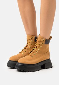 Stivaletti alla caviglia in suede tan con suole in gomma nera, occhielli dorati e lacci arancioni. Presentano un colletto imbottito e un logo sul lato.