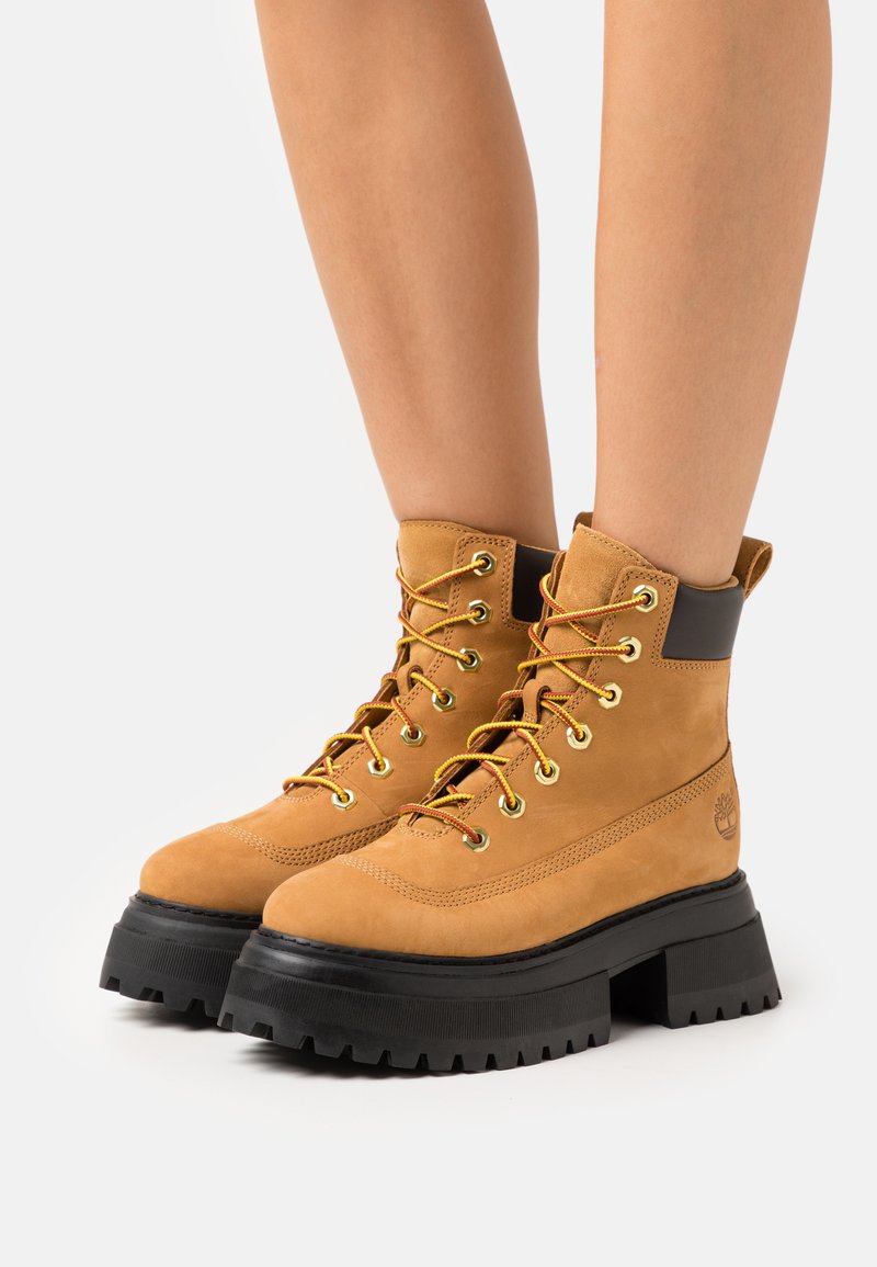Stivaletti alla caviglia in suede tan con suole in gomma nera, occhielli dorati e lacci arancioni. Presentano un colletto imbottito e un logo sul lato.