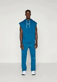 Sweatshirt azul sem mangas com capuz, cordões brancos e logótipo, combinado com calças de treino azuis a condizer com detalhes do logótipo, usado com sapatos brancos.