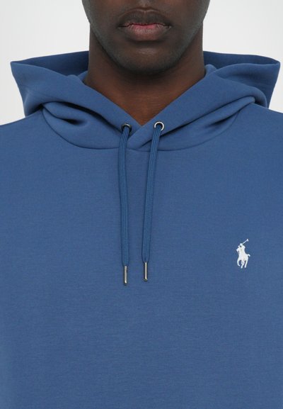 Mėlynas sukapotas džemperis, pagamintas iš minkštos medžiagos, su raišteliais, sidabriniais metaliniais antgaliais ir maža balta logotipu kairėje krūtinės pusėje.