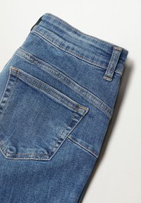 Détail de la poche arrière et de la ceinture d’un jean en denim bleu plié avec des surpiqûres visibles et une boucle de ceinture sur fond blanc.