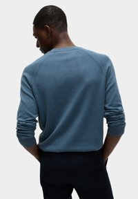 Hellblauer Strickpullover mit rundem Halsausschnitt, gerippten Bündchen und Rückennahtdetails. Schmale Passform, kombiniert mit dunklen Hosen.