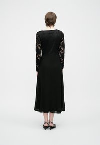 Svart kjole med lange blondermerker, en tettsittende midje og en flytende skjørt. Glatt tekstur med blonderdetaljer og midt på leggen lengde.