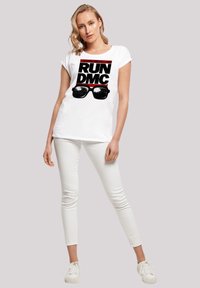 Weißes T-Shirt mit kurzen Ärmeln, das ein großes schwarzes Grafikdesign "RUN DMC" mit Sonnenbrillen zeigt, kombiniert mit passenden weißen Hosen und Sneakern.