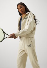 Bežna športna obleka, ki vključuje zip-up kapuco in usklajene trenirke, oboje z logotipom "EA7 Emporio Armani" v črni barvi. Drži teniško lopar.