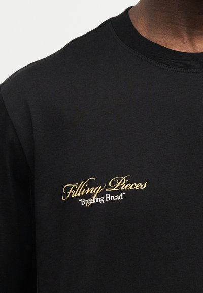 Μαύρο μπλουζάκι με στρογγυλή λαιμόκοψη, με κίτρινη κείμενη γραφή "Filling Pieces" και λευκό κείμενο "Breaking Bread" στο στήθος, φορεμένο από ένα άτομο.
