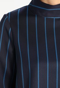 Blusa blu navy con strisce verticali blu, tessuto leggero, colletto elegante e texture liscia. Focus sul dettaglio del motivo a strisce.