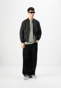 Junger Mann in schwarzer Bomberjacke, olivgrünem T-Shirt, weiten schwarzen Hosen, blau-weißen Sneakers und schwarzen Sonnenbrillen, steht mit einer Hand in der Tasche.