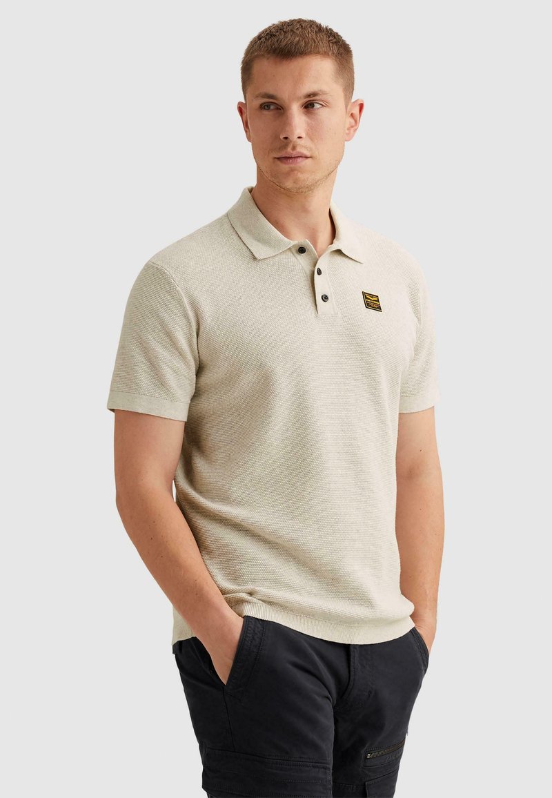 Homme portant un polo beige à manches courtes avec un écusson sur la poitrine et un pantalon noir, debout les mains dans les poches, regardant sur le côté.