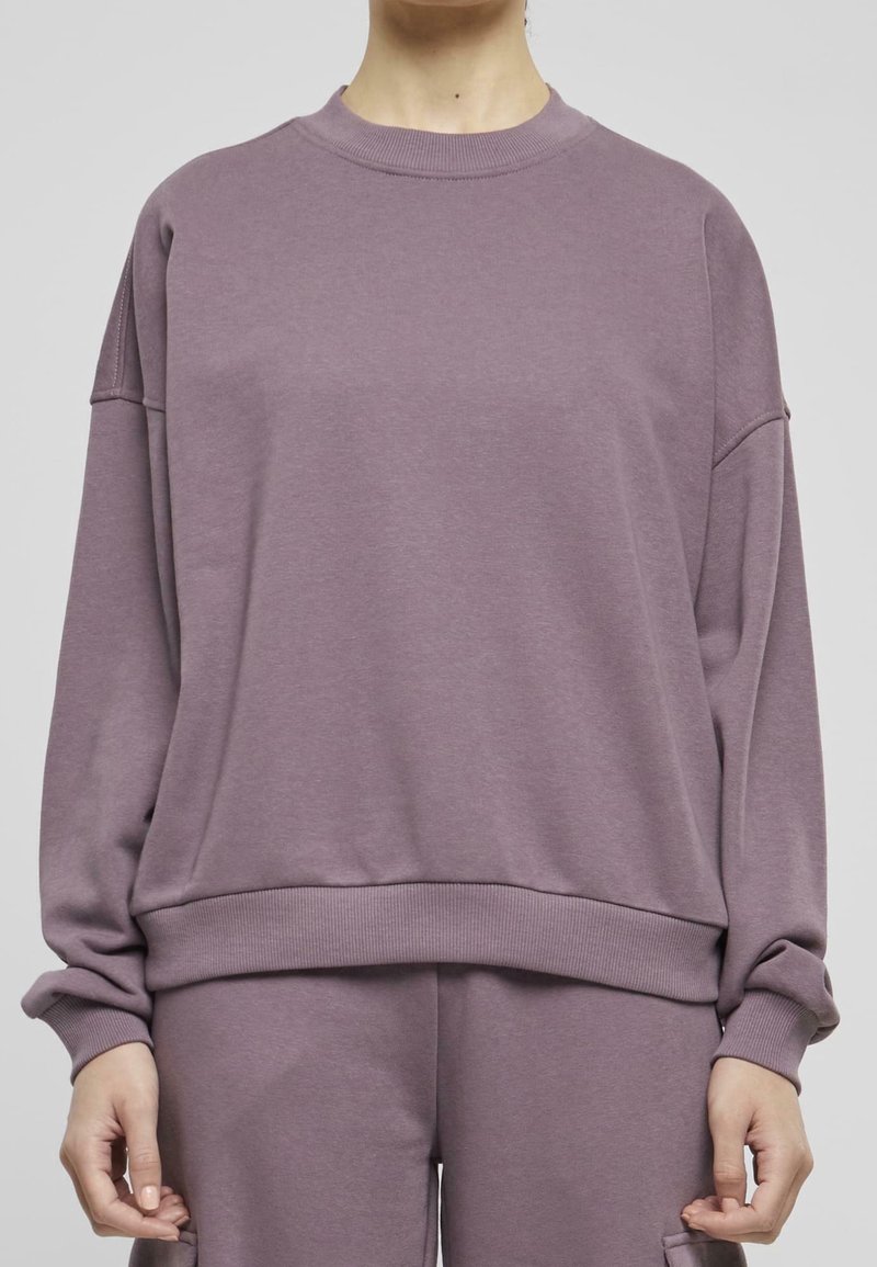 Personne portant un sweat loose mauve et un pantalon assorti, debout devant un fond gris uni.
