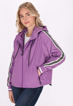 Tânără cu păr blond, purtând o jachetă mov cu glugă, cu dungi alb-negru pe mâneci și blugi bleumarin închisi, cu mâinile în buzunare.