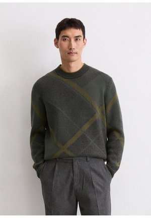 Dark green sweater with a textured pattern featuring diagonal stripes in yellow. Ribbed neckline and cuffs, worn with gray pleated trousers.