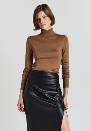 Pull à col roulé marron au design ajusté, à manches longues et avec des poignets côtelés, associé à une jupe en cuir noir avec une fente latérale.