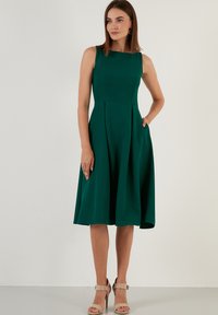 Rochie verde fără mâneci, cu un corp ajustat, fustă plisată și buzunare. Textură netedă, până la genunchi, asortată cu sandale bej cu toc.