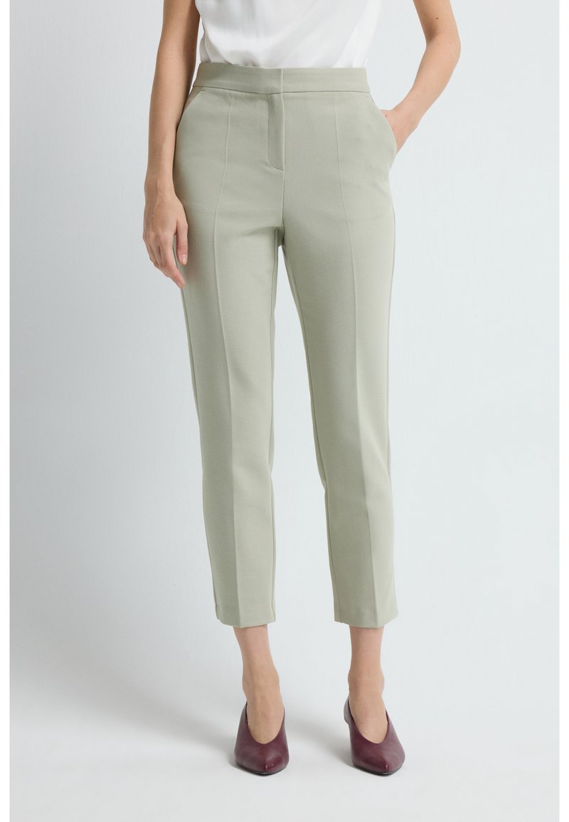 Vrouw in lichtgroene slim-fit enkellange broek, witte top en bordeauxrode puntige hakken, met één hand in haar zak.