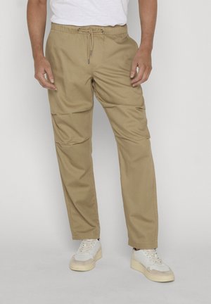 Mężczyzna w beżowych spodniach z krawatem i białych tenisówkach, stojący na tle jasnego, jednolitego tła.