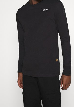 Homme portant un shirt G-Star RAW noir à manches longues et un pantalon cargo noir, debout devant un fond blanc uni.