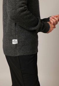 Pull en maille gris foncé avec des poignets et un ourlet côtelés, arborant une petite étiquette de marque blanche sur le côté. Porté par-dessus un pantalon noir.