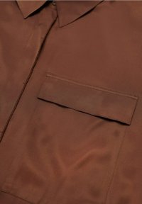 Chemise marron avec poche poitrine gauche à rabat, col visible et patte de boutonnage avant avec boutons dissimulés.