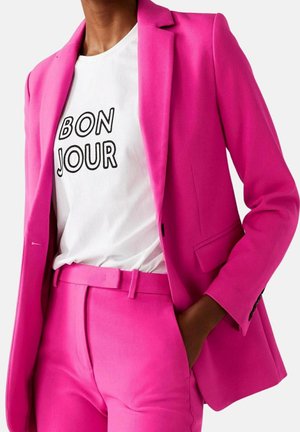 Mujer con traje rosa brillante con blazer y pantalones, combinado con camiseta blanca que muestra el texto "BON JOUR" en letras negras.