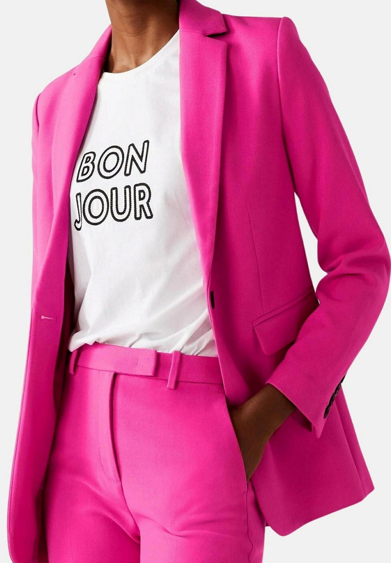 Femme portant un costume rose vif avec blazer et pantalon, associé à un t-shirt blanc affichant le texte "BON JOUR" en lettres noires.