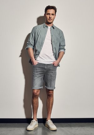 Gestreepte lichtgroene blouse over een wit T-shirt, gecombineerd met opgerolde grijze denimshorts en witte sneakers. Eenvoudig ontwerp en ontspannen pasvorm.