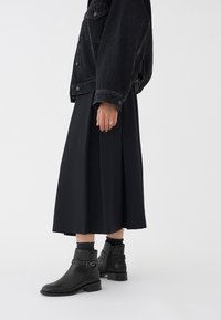 Svart denimjakke over et svart plisserte maxiskjørt, kombinert med svarte lærankleboots med spenne detalj og glidelås på siden.