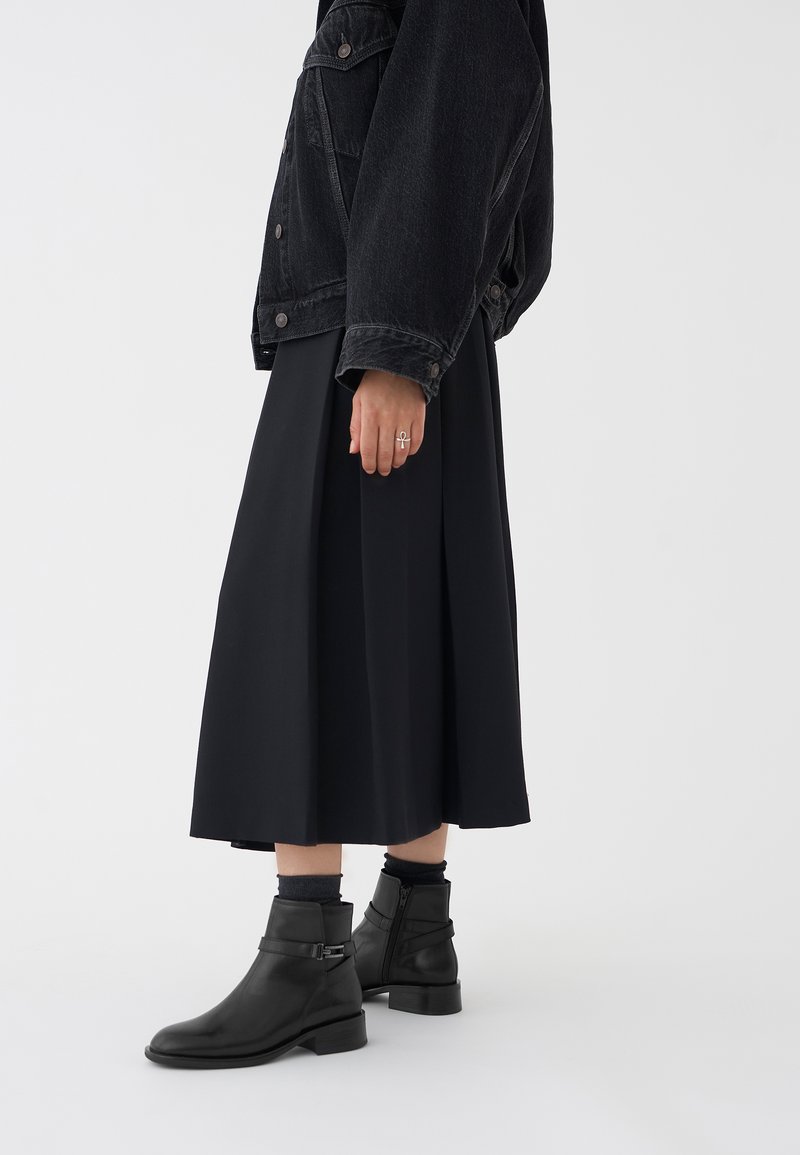Czarna dżinsowa kurtka na czarnej plisowanej maxi spódnicy, zestawiona z czarnymi skórzanymi botkami za kostkę z detalem klamry i bocznym zamkiem błyskawicznym.