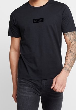 Homme portant un t-shirt Calvin noir et un jean foncé, avec une manchette de tatouage détaillée sur son avant-bras droit.